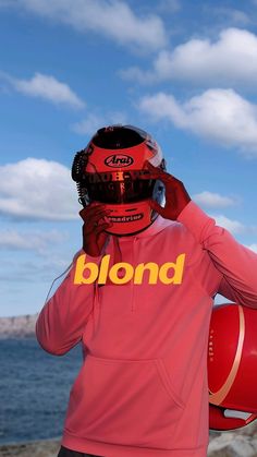 a person wearing a red hoodie and holding a helmet on their head with the ocean in the background