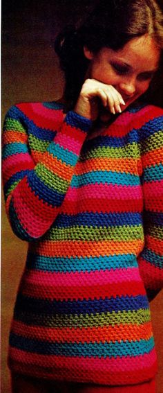 a woman wearing a multicolored sweater and holding her hand to her face while standing in front of a wall