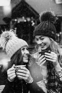 two young women are smiling while looking at their cell phones in the snow, one is holding a cup