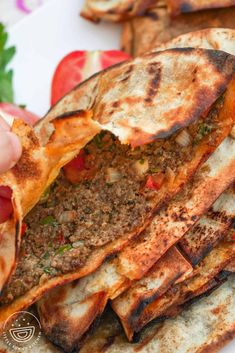 someone is holding up some pita bread with meat and vegetables in the background on a white plate