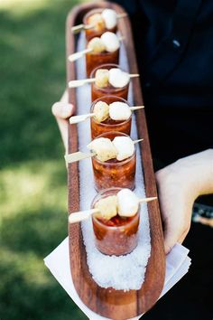 a person holding a long wooden tray filled with small cups and marshmallows