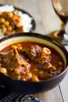 two bowls filled with stew and rice on top of a wooden table next to wine glasses