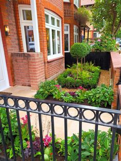 an outdoor garden with flowers and plants on the side of a brick building next to a metal fence