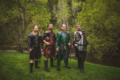 four men dressed in medieval clothing standing next to each other on the grass near a river