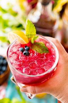 a person holding up a glass filled with blueberries and lemonade on top of a table