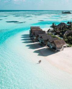 two people walking on the beach in front of some huts and water with palm trees