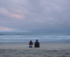 two people sitting on the beach looking out at the ocean with waves in the background