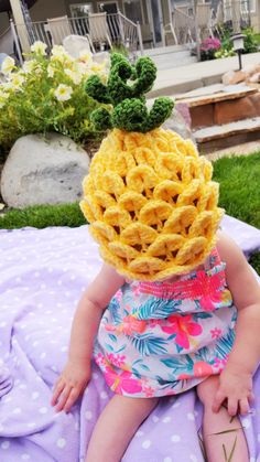 a baby wearing a knitted pineapple hat sitting on top of a purple blanket