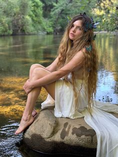 a beautiful young woman sitting on top of a rock next to a body of water