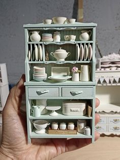 a hand holding a doll house with dishes and cups on it's display shelf