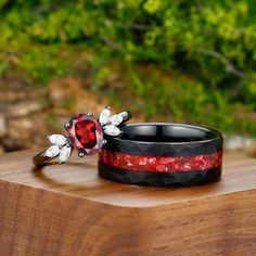 two wedding rings with red and black bands sitting on top of a wooden table next to trees