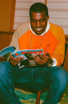 a man sitting on a bed reading a book and smiling at the camera while wearing an orange shirt