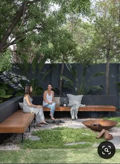 two people sitting on a wooden bench in the middle of a garden with trees and grass