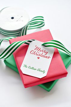 a red and green gift box with a merry christmas tag on it next to a spool of ribbon