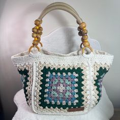 a white purse with green and blue crocheted design on the front, hanging from a wooden handle