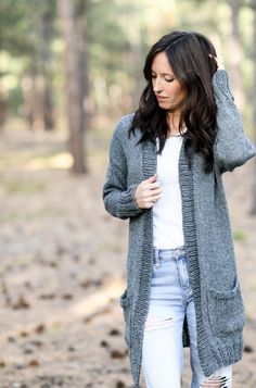 a woman standing in the woods wearing ripped jeans and a gray cardigan sweater with pockets