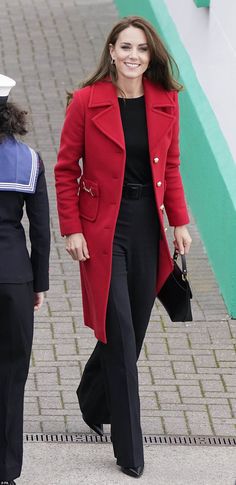 a woman in a red coat and black pants walking down the street with her hand on her hip