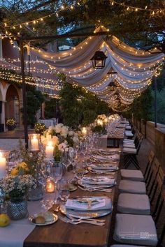 an outdoor dinner table set with candles and flowers on the table, surrounded by string lights