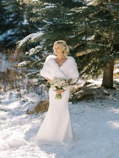 a woman standing in the snow wearing a white dress and fur stole with her hands on her hips