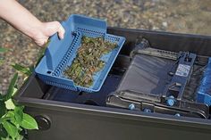 a person holding a plastic container filled with plants and other items in it's trash can