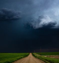 a dirt road in the middle of a green field under a dark cloud filled sky