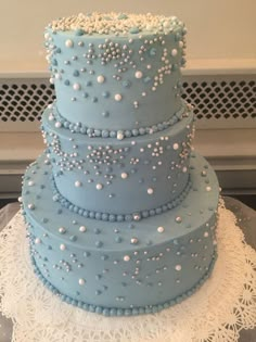 a three tiered blue cake with white pearls on the top and bottom, sitting on a lace doily