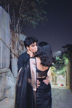 two people standing next to each other in front of a fence and tree at night