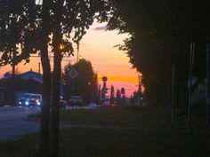 the sun is setting behind some trees and cars on the street in front of them