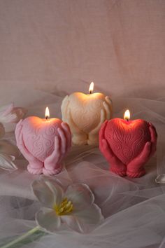 three heart shaped candles sitting next to each other on a white cloth with flowers in the background