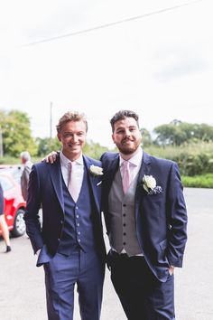 two men in suits standing next to each other on a parking lot with cars behind them