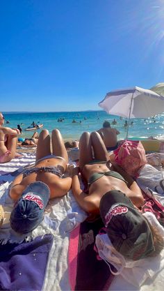 two women laying on the beach in bikinis under an umbrella and sunbathers