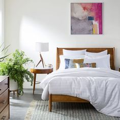 a bed with white sheets and pillows in a bedroom next to a dresser, potted plants and pictures on the wall