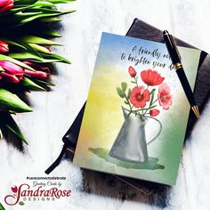 a card with flowers in a silver vase on top of a table next to a bouquet of tulips