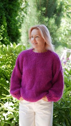 a woman wearing a purple sweater standing in front of some bushes and trees with her hands on her hips