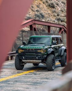 a green jeep driving down a road next to a red metal bridge on a sunny day