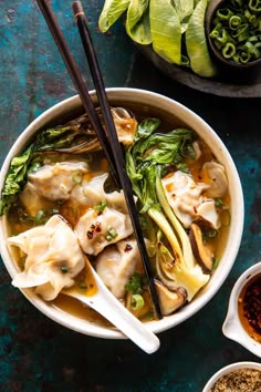 a bowl filled with dumplings and vegetables next to bowls of seasoning on a blue surface