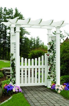 a white arbor with flowers on the ground and in front of it is a brick walkway