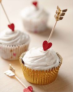 two cupcakes with white frosting and red hearts on top, one has an arrow sticking out of it