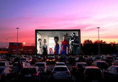 an outdoor movie screen in the middle of a parking lot filled with cars at sunset