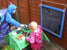 two children in raincoats are playing outside at a picnic table with a sign that says mud pie kitchen