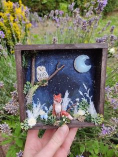 a hand holding up a miniature box with an owl and fox on it in front of flowers