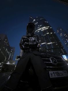 a man sitting on top of a car in front of a tall building at night