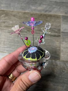 a hand holding a small glass vase with flowers in it's center and two pins sticking out of the top