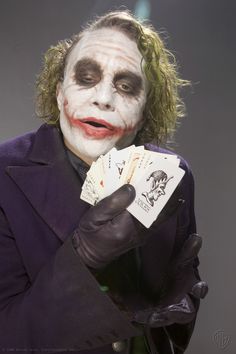 the joker holding playing cards in one hand and wearing a purple suit with white paint on his face