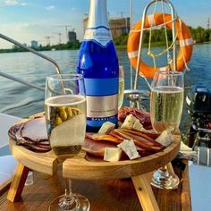 two glasses of wine and cheese on a wooden tray near the water with boats in the background