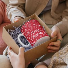 two people sitting on a couch holding a box with a coffee mug in it that says merry christmas