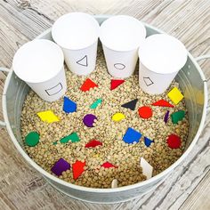 four cups with numbers on them are in a bucket filled with oats and colored shapes