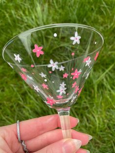 a hand holding a wine glass with pink and white flowers painted on the outside, in front of green grass