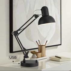 a black lamp on a white desk next to a book and pen holder in front of a painting