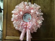 a pink wreath hanging from the side of a wooden door with ribbon around it and a clock in the center